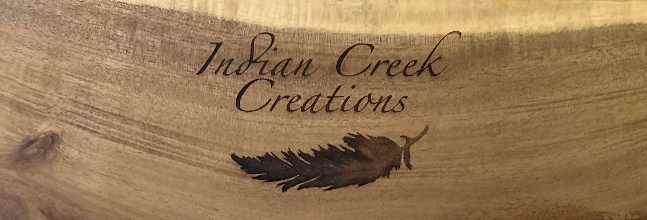 Wooden cutting board custom engraved with 'Indian Creek Creations' branding on a stone surface with greenery. Personalized laser engraving. 